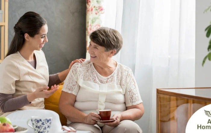 Smiling caregiver with her hand on an older woman’s shoulder as they share tea on the couch, showing home care vs assisted living